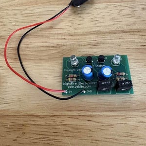 May include: A green electronic circuit board labeled NightFire Electronics LED Flasher with two clear LEDs, two blue capacitors, four resistors, and two black transistors. A black 9V battery connector with red and black wires is attached.