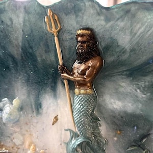 May include: A decorative resin art piece featuring a bronze-toned merman figure with a beard and crown, holding a gold trident. The figure is set against a swirling blue and white ocean wave background with embedded seashells and sand textures.