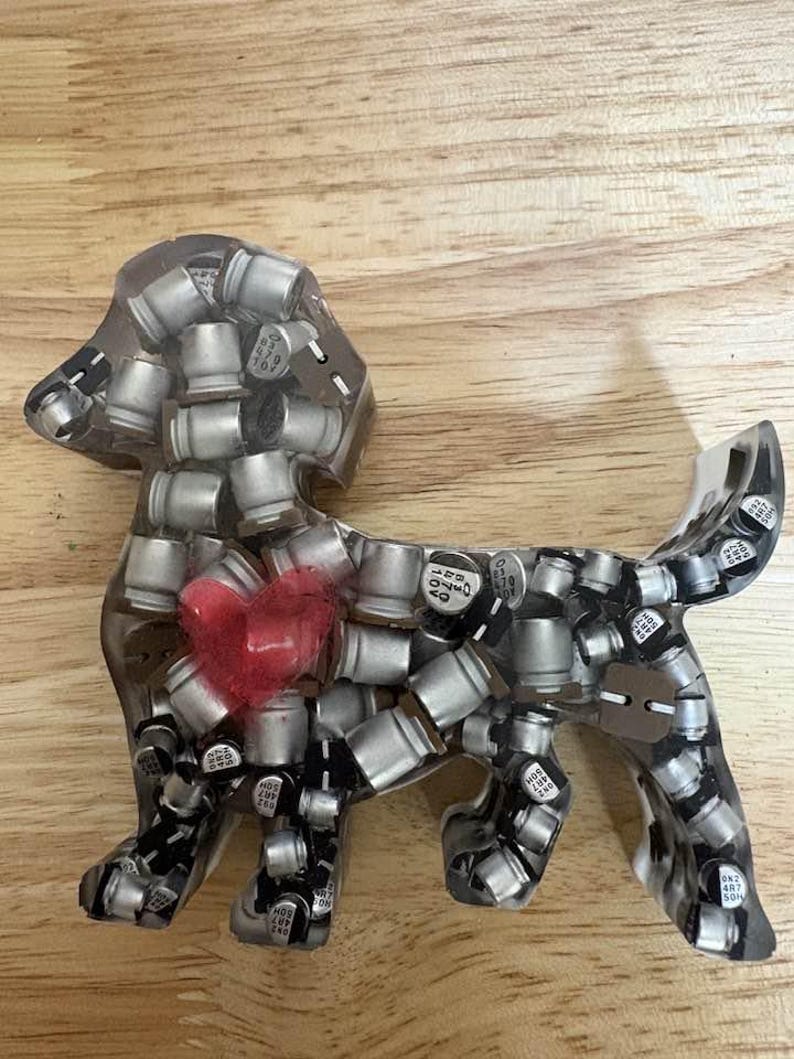 May include: A clear resin dog figurine filled with silver electronic components. A red heart is embedded in the center of the dog's body. The figurine is displayed on a wooden surface.