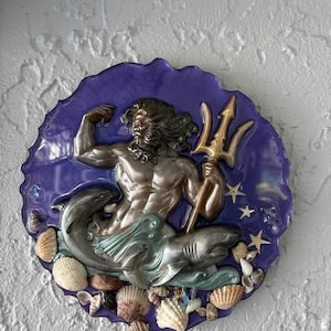 May include: A circular wall plaque featuring a metallic bronze-toned figure of Poseidon holding a trident, surrounded by a deep purple resin background. The base is decorated with various natural seashells and a sculpted shark figure.