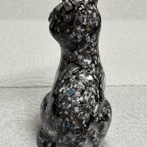 Puede incluir: Figura decorativa de gato con un patr&oacute;n de mosaico de peque&ntilde;as piezas rectangulares en tonos negro, blanco y gris. El gato est&aacute; sentado, con la cabeza ligeramente girada hacia un lado. La figura tiene un acabado brillante.