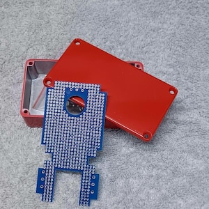 May include: A red plastic project box with a matching lid and a blue circuit board. The box is open, revealing the interior. The circuit board has numerous holes for components.
