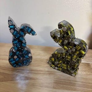 K&ouml;nnte beinhalten: Zwei abstrakte Skulpturen, eine blaue und eine gelbe, aus kleinen zylindrischen Objekten, die in klarem Harz eingeschlossen sind. Die blaue Skulptur ist eine stilisierte Form, w&auml;hrend die gelbe eine sitzende Form ist. Beide stehen auf einer Holzoberfl&auml;che.