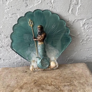 May include: A decorative resin shell-shaped plaque featuring a dark-skinned merman with a gold crown and trident. The merman has a teal-colored tail and is positioned against a translucent teal shell background with small seashells at the base.