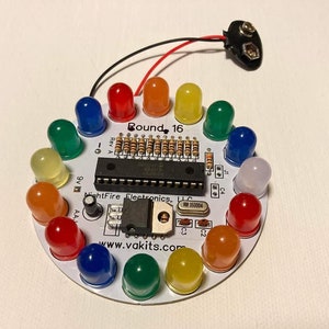 May include: A white circuit board with a round shape and 16 LEDs in a circle. The LEDs are red, orange, yellow, green, blue, and white. The board has the text "Round 16" printed on it.