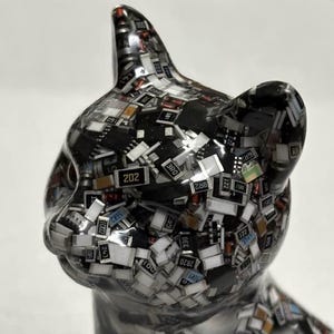 Puede incluir: Una brillante figura de gato plateado, cuya superficie est&aacute; cubierta por un mosaico de diminutos componentes electr&oacute;nicos. Los componentes son negros, blancos y de varios colores, creando una apariencia texturizada &uacute;nica. Las orejas del gato est&aacute;n erguidas y sus ojos est&aacute;n cerrados.