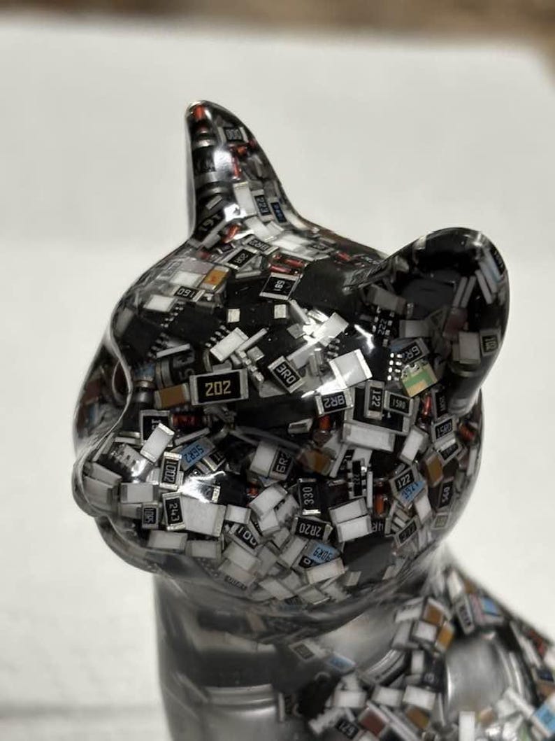 Puede incluir: Una brillante figura de gato plateado, cuya superficie est&aacute; cubierta por un mosaico de diminutos componentes electr&oacute;nicos. Los componentes son negros, blancos y de varios colores, creando una apariencia texturizada &uacute;nica. Las orejas del gato est&aacute;n erguidas y sus ojos est&aacute;n cerrados.