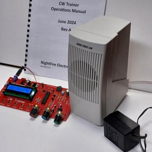 May include: A red circuit board with the text "CW Trainer" and several knobs and switches. The board is connected to a white speaker with the text "SOUND FORCE 540" and a black power adapter. A manual titled "CW Trainer Operations Manual June 2024 Rev A" is visible in the background.