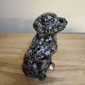 May include: A ceramic dog figurine in a sitting position. The dog is black and white, with a mosaic-like pattern. A small, silver-colored tag is on the dog's chest. The figurine is on a wooden surface.