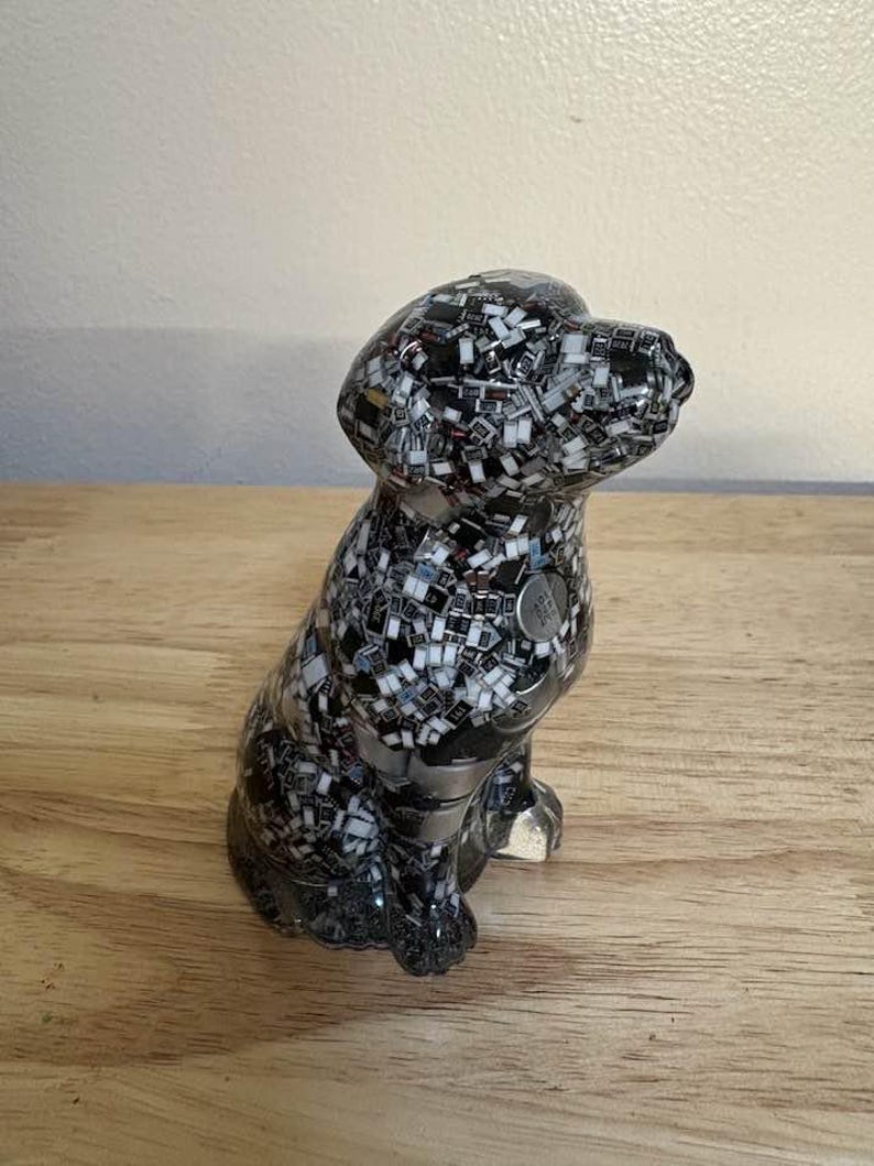 May include: A ceramic dog figurine in a sitting position. The dog is black and white, with a mosaic-like pattern. A small, silver-colored tag is on the dog's chest. The figurine is on a wooden surface.