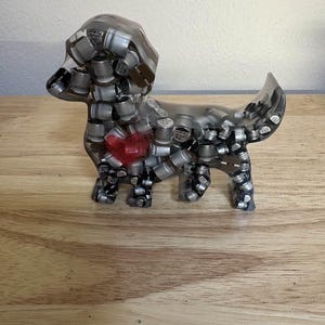 May include: A clear resin dachshund sculpture filled with silver and black cylindrical components. A red heart shape is embedded in the dog's chest. The sculpture is displayed on a wooden surface.