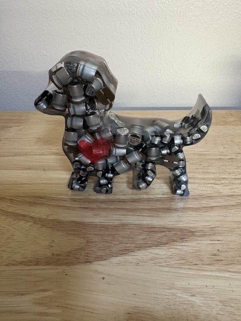 May include: A clear resin dachshund sculpture filled with silver and black cylindrical components. A red heart shape is embedded in the dog's chest. The sculpture is displayed on a wooden surface.
