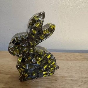 K&ouml;nnte beinhalten: Eine transparente Hasenfigur aus Harz, gef&uuml;llt mit kleinen, zylindrischen Objekten mit silbernen Deckeln und gelben Innenr&auml;umen. Der Hase ist aufrecht positioniert, mit erhobenen Ohren. Die Figur wird auf einer Holzoberfl&auml;che ausgestellt.