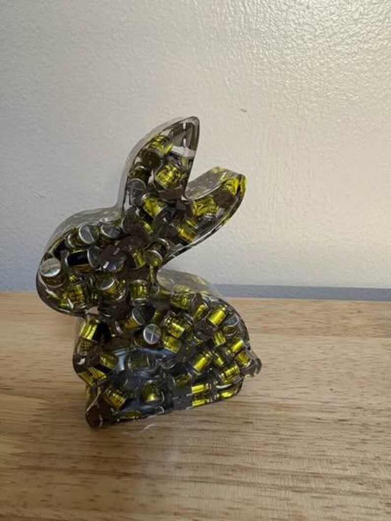 K&ouml;nnte beinhalten: Eine transparente Hasenfigur aus Harz, gef&uuml;llt mit kleinen, zylindrischen Objekten mit silbernen Deckeln und gelben Innenr&auml;umen. Der Hase ist aufrecht positioniert, mit erhobenen Ohren. Die Figur wird auf einer Holzoberfl&auml;che ausgestellt.