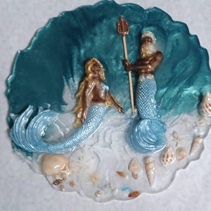 May include: A resin art piece featuring a mermaid and a merman with a trident, set against a teal wave backdrop. The base includes seashells and sand-like details, creating a coastal theme. The figures have gold and blue accents.