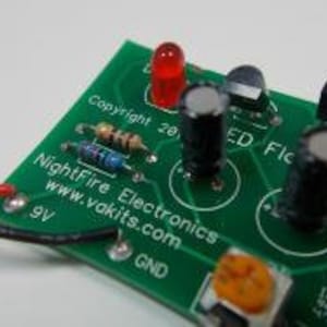 May include: A green circuit board with a red LED, a black resistor, a black capacitor, and a black switch. The board has the text "Nightfire Electronics" and "www.yakits.com" printed on it. The board also has the text "Copyright 2009" printed on it.