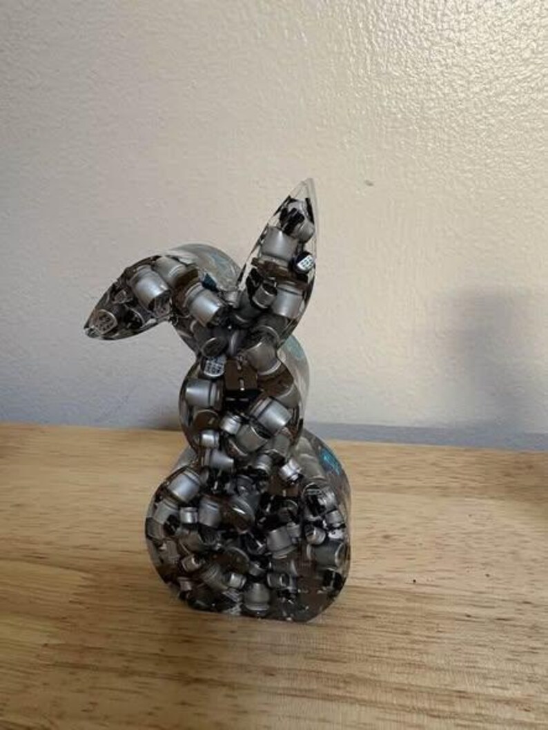 K&ouml;nnte beinhalten: Eine klare Harzskulptur eines Hasen, gef&uuml;llt mit kleinen, silberfarbenen zylindrischen Objekten. Der Hase hat aufrechte Ohren und einen abgerundeten K&ouml;rper. Die Skulptur wird auf einer Holzoberfl&auml;che vor einer neutralen Wand ausgestellt.