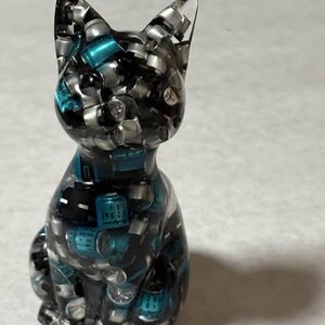 Puede incluir: Una figura de gato de resina transparente llena de peque&ntilde;os componentes plateados y verde azulado. El gato est&aacute; sentado erguido con las orejas levantadas. La figura tiene un acabado brillante y est&aacute; sobre un fondo neutro.