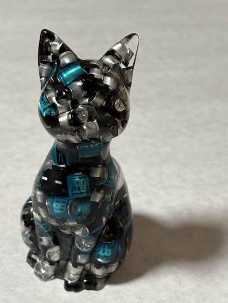 Puede incluir: Una figura de gato de resina transparente llena de peque&ntilde;os componentes plateados y verde azulado. El gato est&aacute; sentado erguido con las orejas levantadas. La figura tiene un acabado brillante y est&aacute; sobre un fondo neutro.
