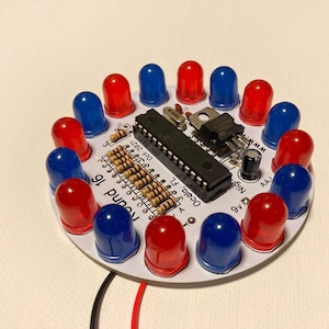 May include: A round circuit board with a microcontroller, resistors, and a 9V battery connector. The board has 16 LEDs arranged in a circle, alternating red and blue.