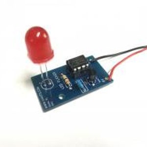 May include: A red LED light is connected to a small blue circuit board with a black and red wire. The circuit board has a resistor and an integrated circuit chip.