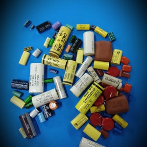 May include: A collection of various electronic components, including capacitors, resistors, and transistors. The components are in a variety of colors, including yellow, red, blue, and green. Some of the components have text printed on them, such as "WMF 05W2", "2.0 MFD", and "100 VDC".