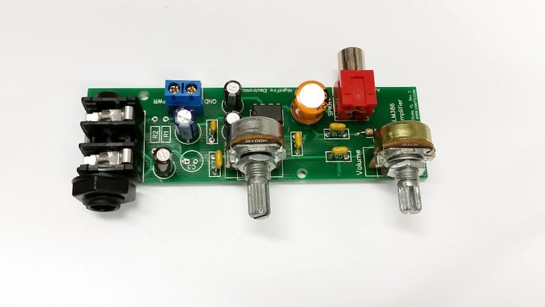 Puede incluir: Una placa de circuito verde con una toma de audio negra, un bloque de terminales azul y rojo, tres potenci&oacute;metros y un conector RCA rojo y negro. La placa tiene el texto "LM386 Amplifier" impreso en ella.