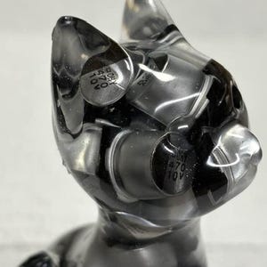 Puede incluir: Una escultura transparente con forma de gato con componentes negros y plateados incrustados. La escultura tiene un acabado liso y brillante y un dise&ntilde;o estilizado. Los componentes parecen ser piezas electr&oacute;nicas, lo que a&ntilde;ade una est&eacute;tica industrial &uacute;nica a la obra de arte.