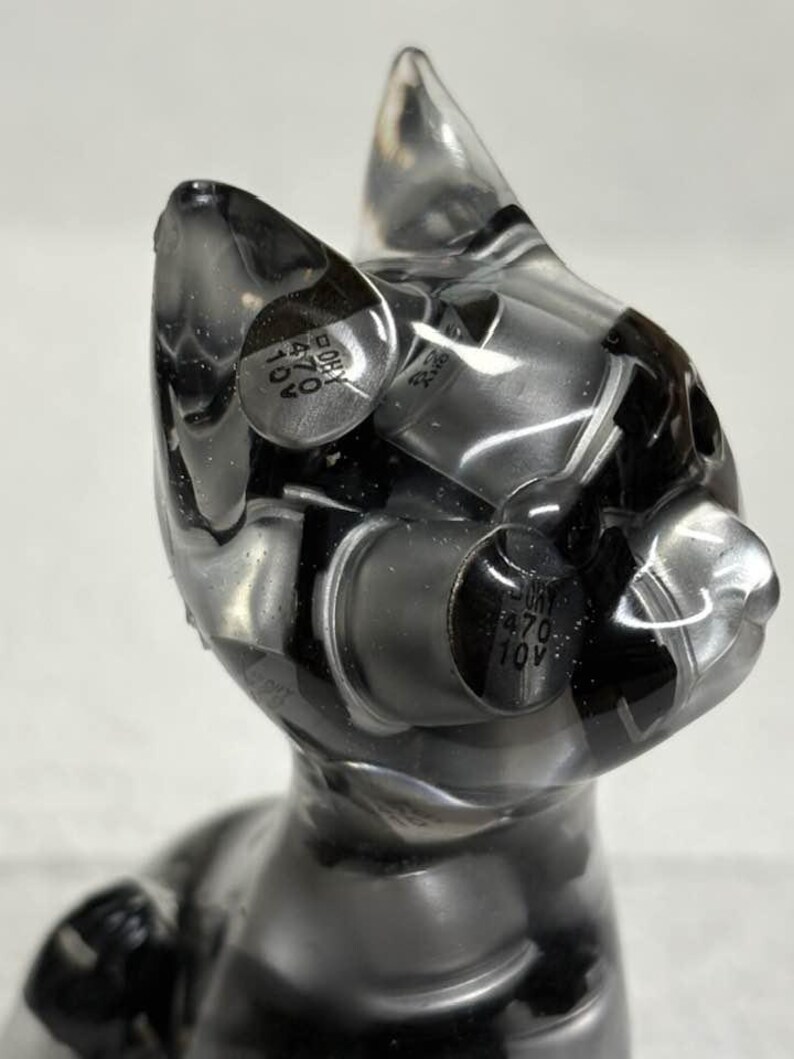 Puede incluir: Una escultura transparente con forma de gato con componentes negros y plateados incrustados. La escultura tiene un acabado liso y brillante y un dise&ntilde;o estilizado. Los componentes parecen ser piezas electr&oacute;nicas, lo que a&ntilde;ade una est&eacute;tica industrial &uacute;nica a la obra de arte.