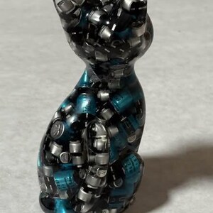 Puede incluir: Una figura de gato de resina con un dise&ntilde;o &uacute;nico. El gato est&aacute; compuesto por peque&ntilde;os componentes plateados y resina de color turquesa, creando un efecto mosaico. El gato est&aacute; en posici&oacute;n sentada, con las orejas erguidas.