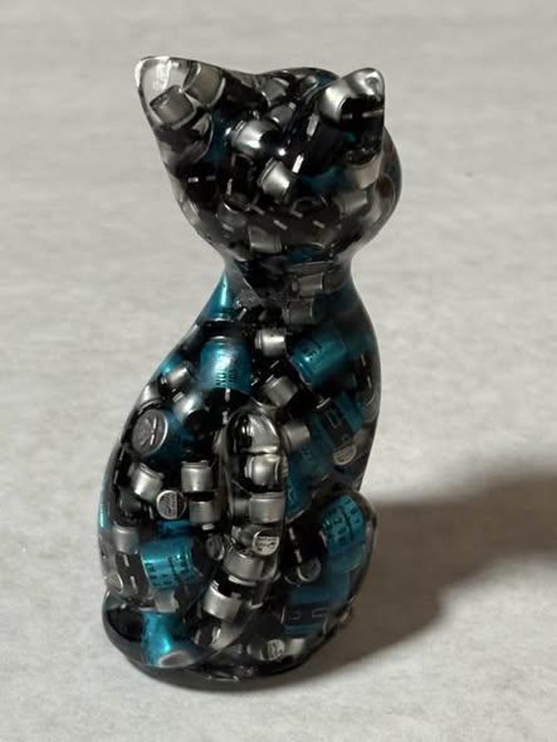 Puede incluir: Una figura de gato de resina con un dise&ntilde;o &uacute;nico. El gato est&aacute; compuesto por peque&ntilde;os componentes plateados y resina de color turquesa, creando un efecto mosaico. El gato est&aacute; en posici&oacute;n sentada, con las orejas erguidas.