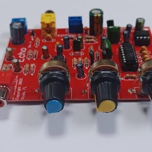 May include: A red circuit board with various electronic components, including potentiometers with blue, yellow, and white knobs. The board has the word "echo" printed on it, along with other text. A red RCA connector is visible on the left side.