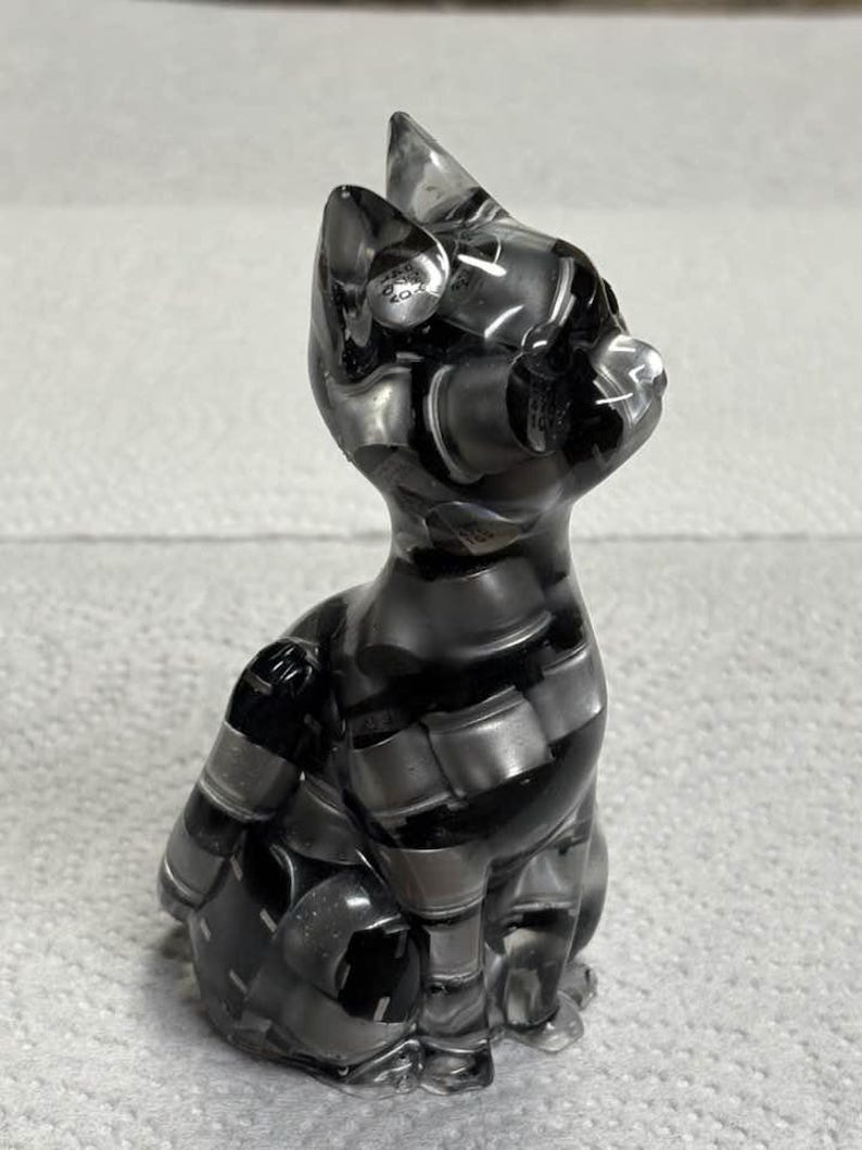 Puede incluir: Una figura de gato gris transparente con un patr&oacute;n de rayas. El gato est&aacute; sentado, con las orejas erguidas. La figura est&aacute; hecha de un material transparente, que permite el paso de la luz.