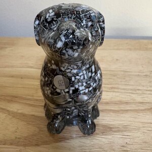 May include: A decorative dog figurine made from a mosaic of small, black, white, and gray electronic components. The dog is in a sitting position with a small, round, silver-colored emblem on its side.