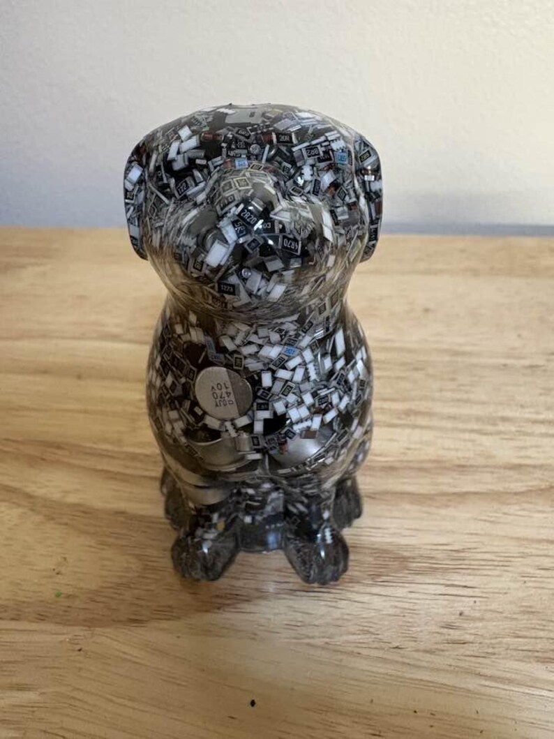 May include: A decorative dog figurine made from a mosaic of small, black, white, and gray electronic components. The dog is in a sitting position with a small, round, silver-colored emblem on its side.