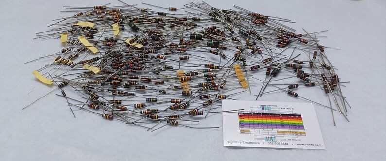 May include: A large assortment of resistors with color bands, spread out on a white surface. The resistors are arranged in a random pattern, with some overlapping. A small card with a resistor color code chart is visible in the lower right corner of the image.