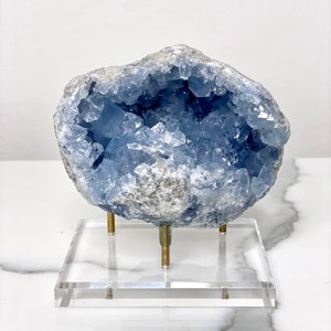May include: A blue celestite crystal geode on a clear acrylic stand with gold metal legs. The geode is a natural formation with a hollow center lined with crystals.