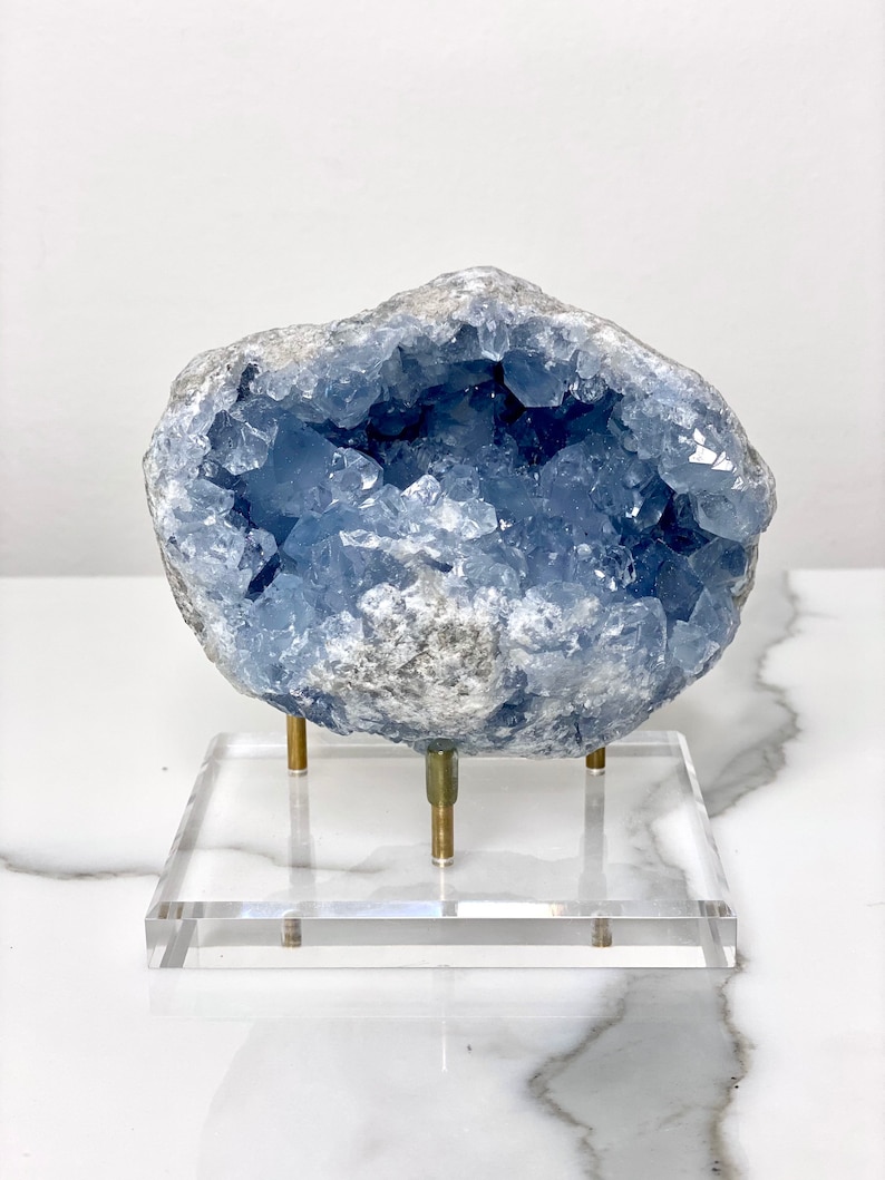 May include: A blue celestite crystal geode on a clear acrylic stand with gold metal legs. The geode is a natural formation with a hollow center lined with crystals.