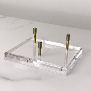 May include: A clear acrylic display stand with three brass posts. The stand is rectangular and has a smooth surface.