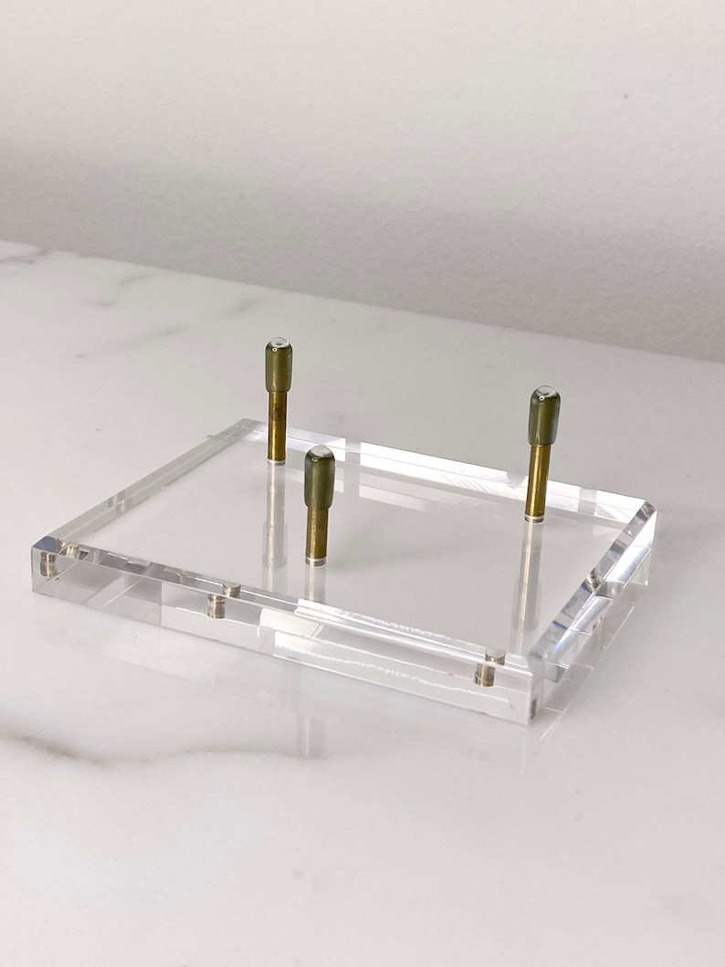 May include: A clear acrylic display stand with three brass posts. The stand is rectangular and has a smooth surface.