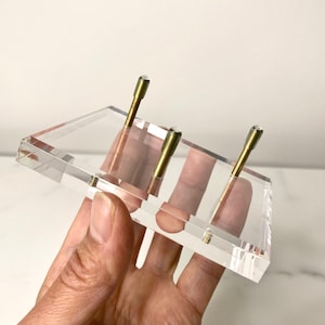 May include: A clear acrylic display stand with three brass posts. The stand is designed to hold small items, such as jewelry or figurines.