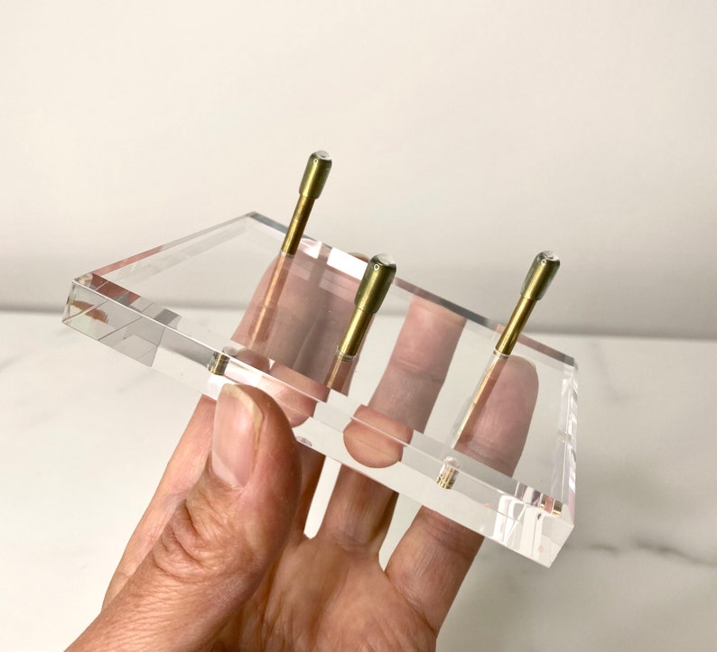 May include: A clear acrylic display stand with three brass posts. The stand is designed to hold small items, such as jewelry or figurines.