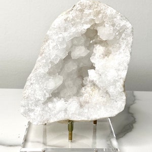 May include: A large white crystal geode with a natural opening revealing a cluster of smaller crystals inside. The geode is displayed on a clear acrylic stand with brass accents.
