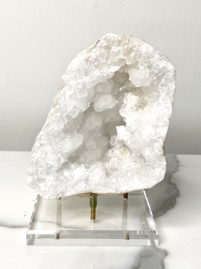 May include: A large white crystal geode with a natural opening revealing a cluster of smaller crystals inside. The geode is displayed on a clear acrylic stand with brass accents.