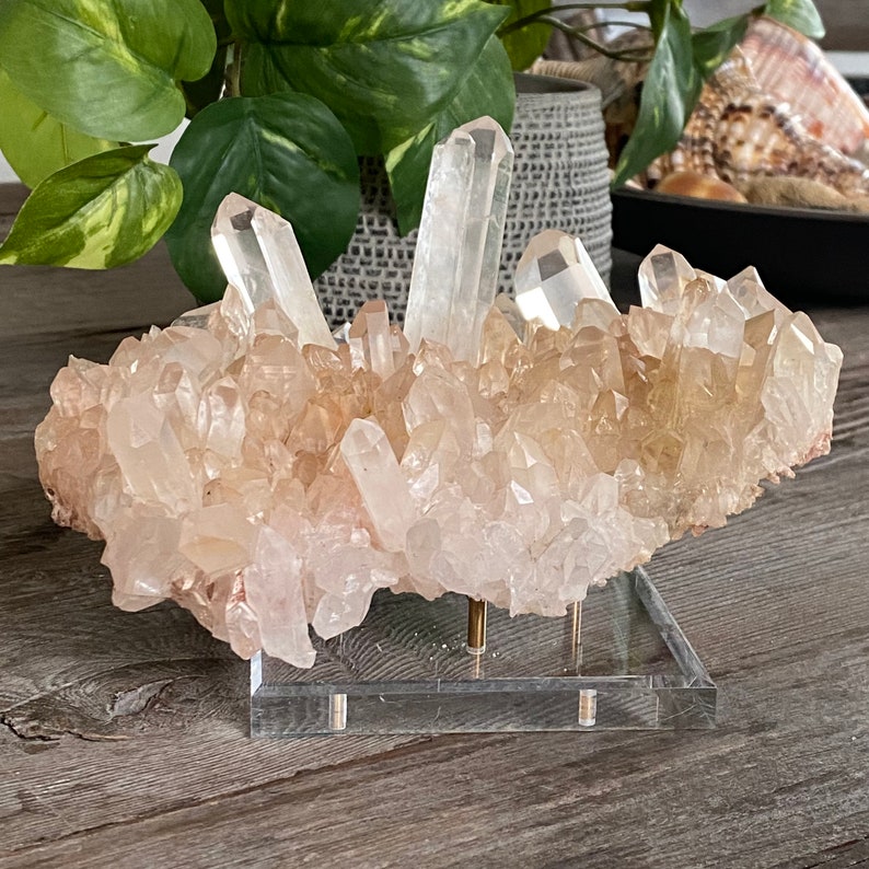 May include: A large cluster of clear quartz crystals with a pink hue, displayed on a clear acrylic stand. The crystals are arranged in a natural, organic shape, with some of the points reaching upwards.