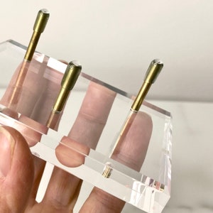 May include: Three gold-colored metal rods with clear glass tops are displayed on a clear acrylic stand. The stand is held by a hand.