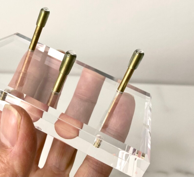 May include: Three gold-colored metal rods with clear glass tops are displayed on a clear acrylic stand. The stand is held by a hand.
