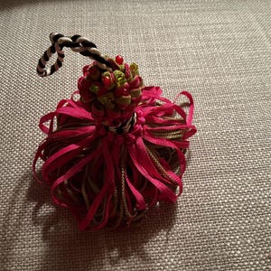 Puede incluir: Una borla decorativa con un lazo para colgar. La borla presenta un grupo de elementos rojos, verdes y dorados en la parte superior, con una cascada de cintas rojas que forman el cuerpo principal. El lazo para colgar es blanco y negro.