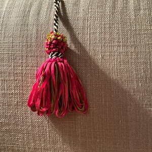 Puede incluir: Una borla decorativa con un cord&oacute;n trenzado en blanco y negro, una secci&oacute;n superior tejida con detalles verdes y rosas, y una falda de cintas rosa brillante. La borla se encuentra sobre un fondo neutro y texturizado.