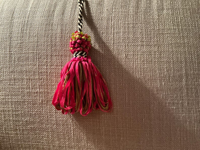 Puede incluir: Una borla decorativa con un cord&oacute;n trenzado en blanco y negro, una secci&oacute;n superior tejida con detalles verdes y rosas, y una falda de cintas rosa brillante. La borla se encuentra sobre un fondo neutro y texturizado.